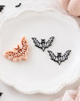 a white plate with three black bat-shaped cookie cutters, two of which are black and one is orange, placed on a white surface. There are also two small pumpkins and some black bats scattered around the plate.