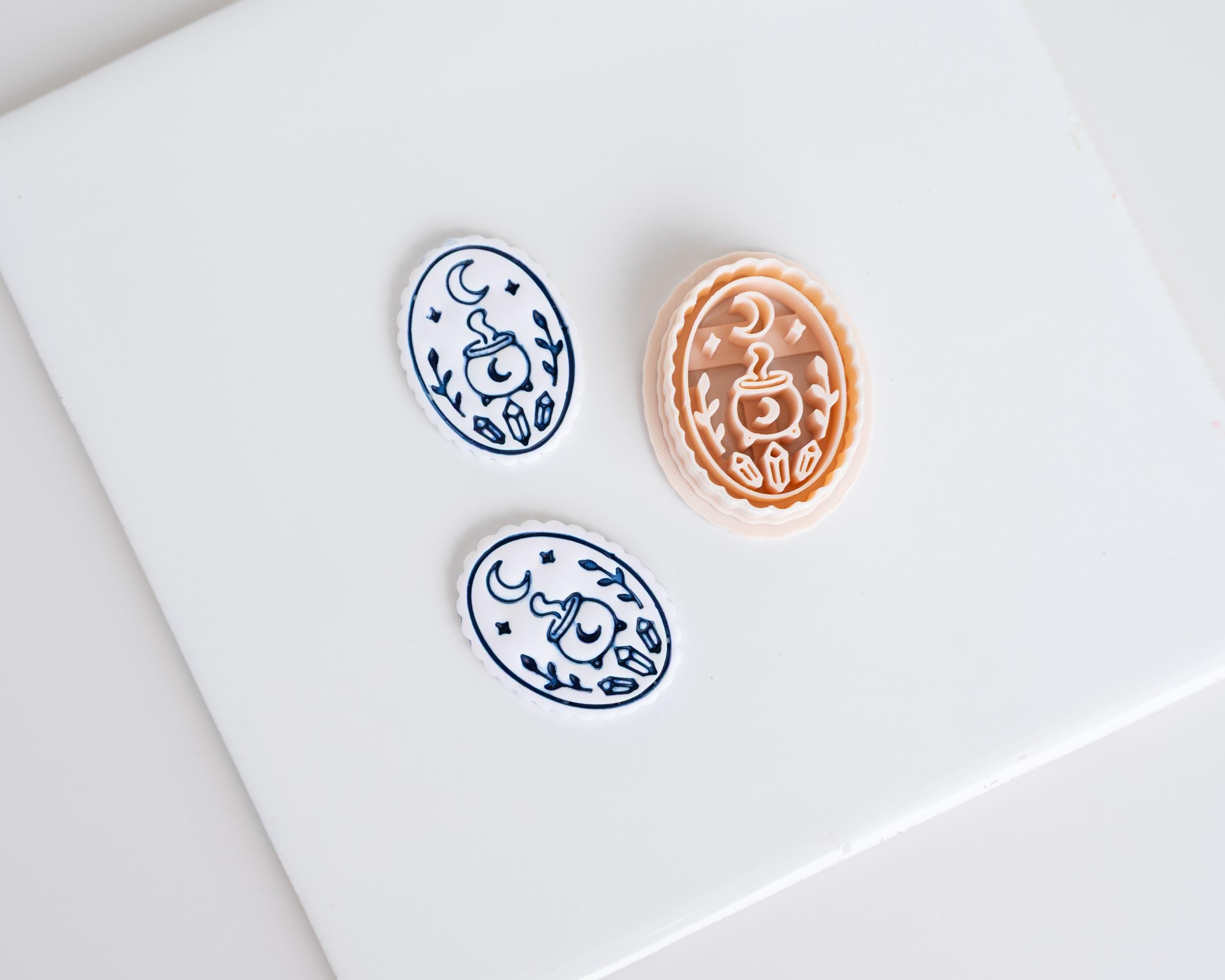 Halloween clay earring cutter featuring a crescent moon, steaming cauldron, crystals and foliage with two matching blue and white finished pieces.