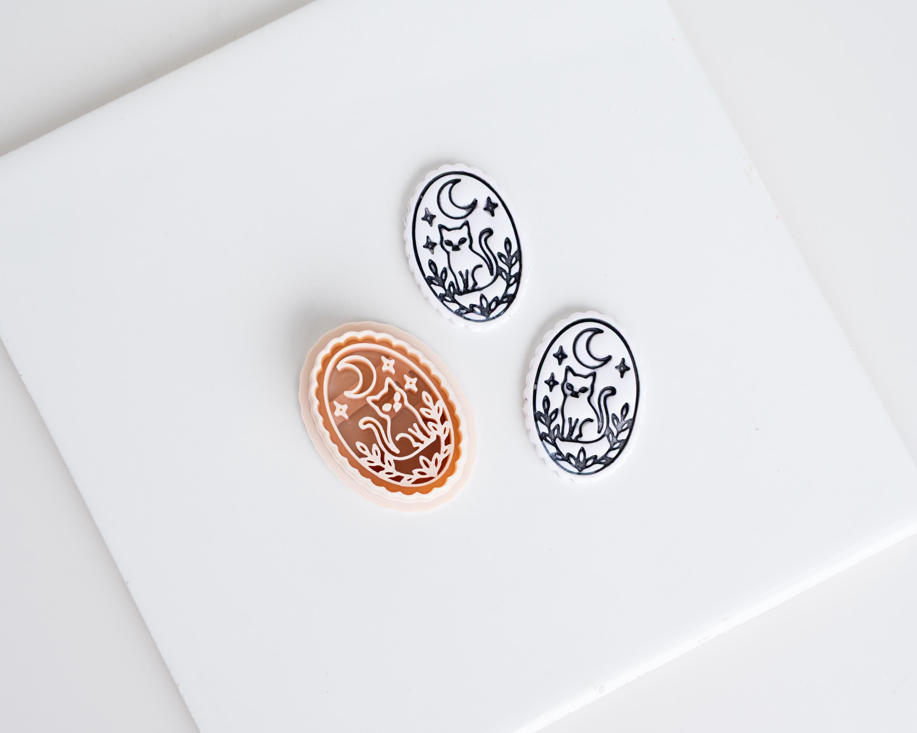 Halloween oval clay cutter with crescent moon and cat design shown beside two matching black and white polymer clay earring pieces.