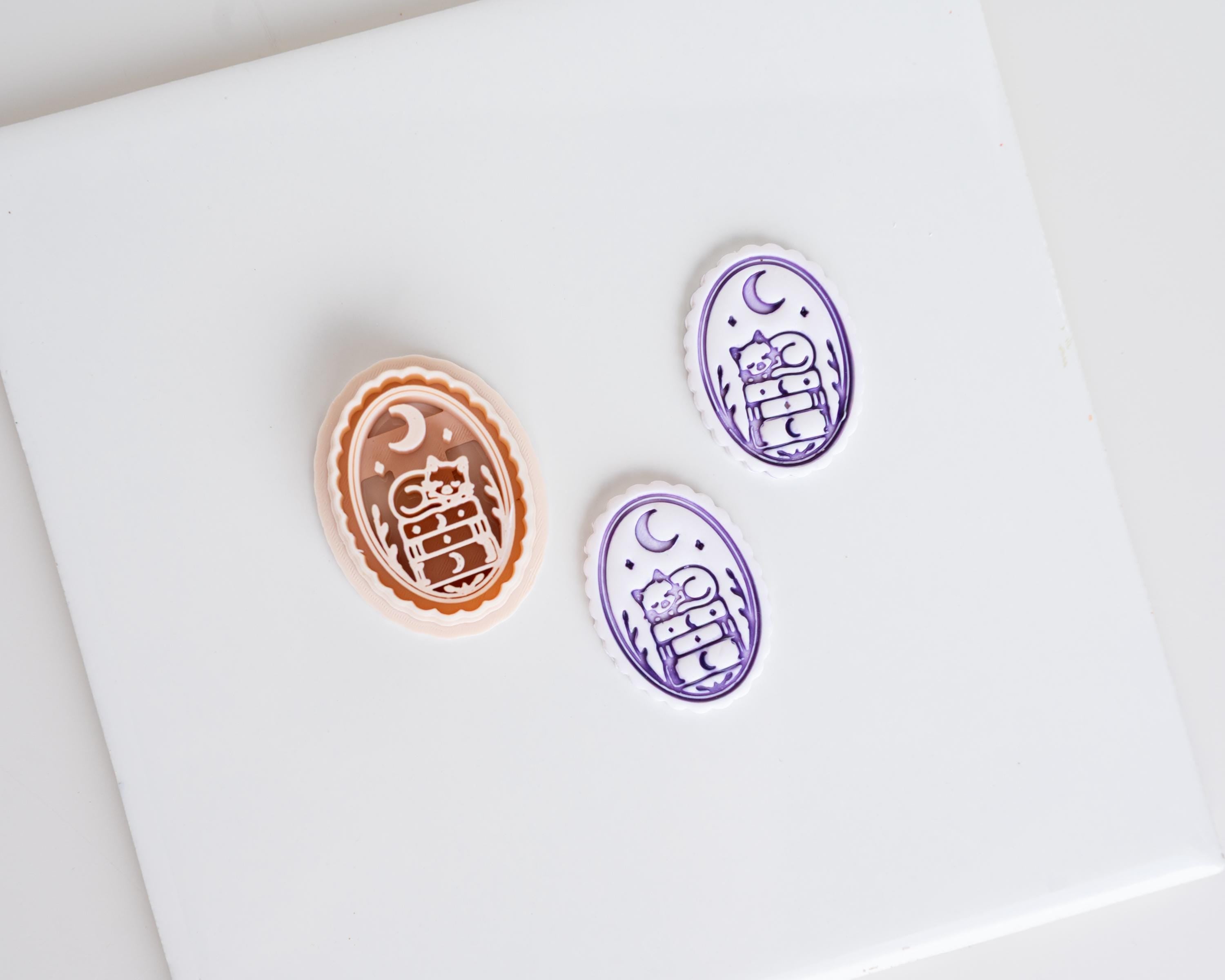Halloween clay cutter featuring a cat resting on stacked books beneath a crescent moon with two matching purple polymer clay earring pieces.