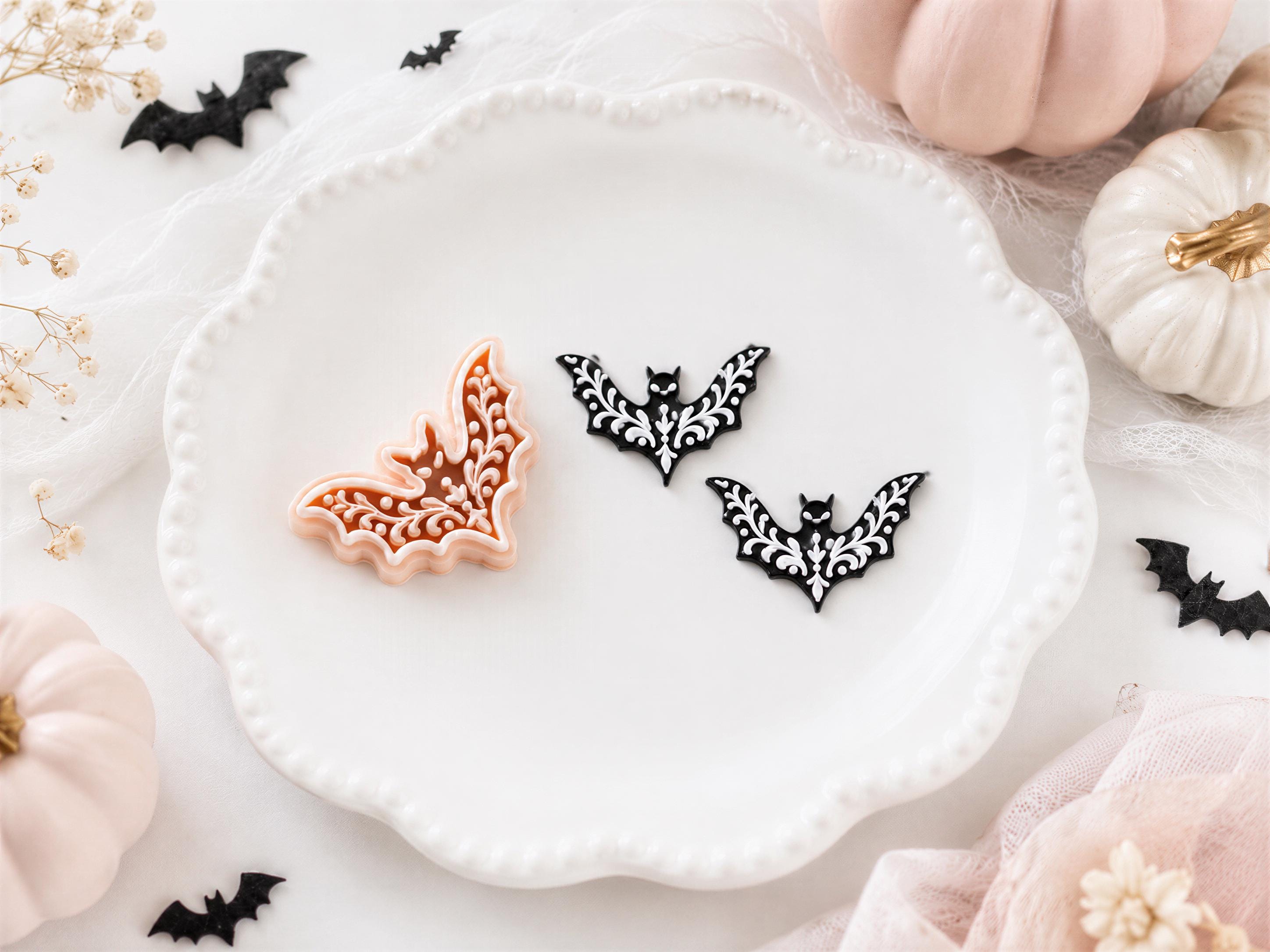 a white plate with three black bat-shaped cookie cutters, two of which are black and one is orange, placed on a white surface. There are also two small pumpkins and some black bats scattered around the plate.