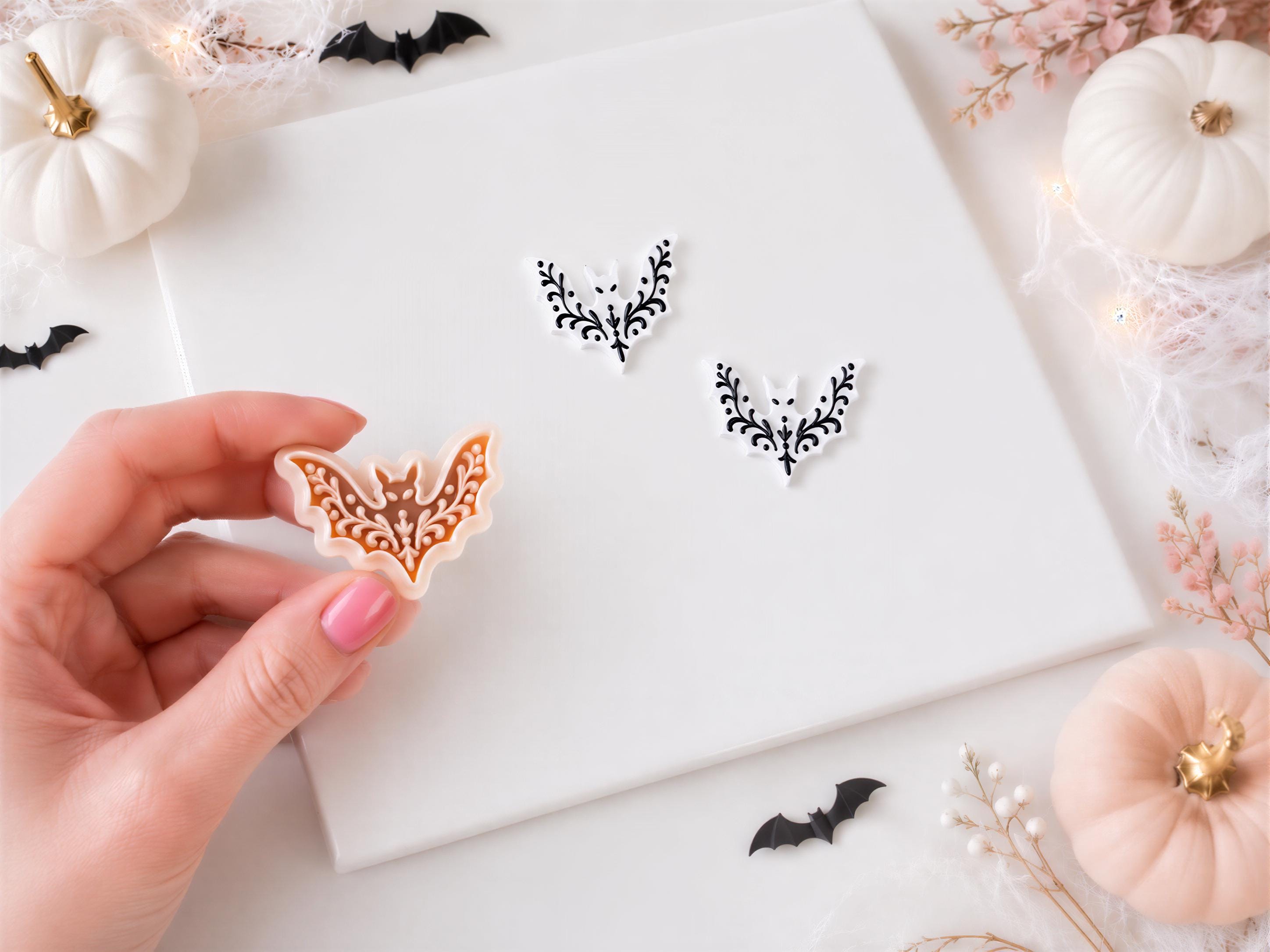 A hand holding a bat-shaped cookie cutter, surrounded by Halloween decorations such as pumpkins, bats, and floral accents on a white background.