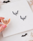 A hand holding a bat-shaped cookie cutter, surrounded by Halloween decorations such as pumpkins, bats, and floral accents on a white background.